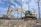 An excavator is actively working on ground excavation in a barren landscape with scattered dry trees and a clear blue sky in the background. The construction equipment is yellow with visible tracks, and a person is operating it from inside the cabin.