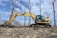 An excavator is actively working on ground excavation in a barren landscape with scattered dry trees and a clear blue sky in the background. The construction equipment is yellow with visible tracks, and a person is operating it from inside the cabin.