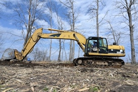 An excavator is actively working on ground excavation in a barren landscape with scattered dry trees and a clear blue sky in the background. The construction equipment is yellow with visible tracks, and a person is operating it from inside the cabin.