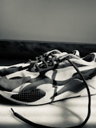 A monochrome sneaker with a modern design featuring curved patterns and textured surfaces. The sneaker is placed on a light-colored floor, with one of the laces extending outwards. The lighting creates strong shadows and highlights the shoe's contours.