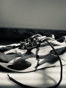 A monochrome sneaker with a modern design featuring curved patterns and textured surfaces. The sneaker is placed on a light-colored floor, with one of the laces extending outwards. The lighting creates strong shadows and highlights the shoe's contours.