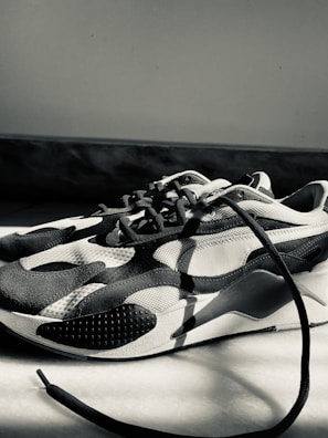A monochrome sneaker with a modern design featuring curved patterns and textured surfaces. The sneaker is placed on a light-colored floor, with one of the laces extending outwards. The lighting creates strong shadows and highlights the shoe's contours.