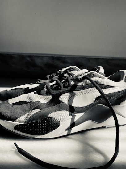 A monochrome sneaker with a modern design featuring curved patterns and textured surfaces. The sneaker is placed on a light-colored floor, with one of the laces extending outwards. The lighting creates strong shadows and highlights the shoe's contours.