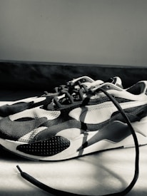 A monochrome sneaker with a modern design featuring curved patterns and textured surfaces. The sneaker is placed on a light-colored floor, with one of the laces extending outwards. The lighting creates strong shadows and highlights the shoe's contours.