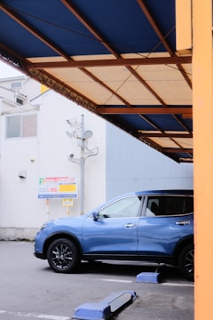 A blue SUV is parked in a designated parking space. The surrounding area includes a white building in the background and a covered structure with a blue and beige awning. A parking lot sign with pricing information is visible.