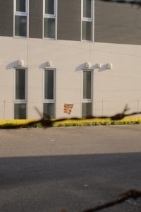 A multi-story building exterior with modern design features elongated vertical windows and two-tone wall paneling. Shadows cast by the sun create contrast, while a small, colorful sign hangs near one of the windows. In the foreground, a wire fence partially obstructs the view.