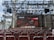 A stage setup with a large screen displaying logos for ASUS and Republic of Gamers. Rows of red chairs are arranged in front of the stage. The event is titled 'DEVFEST BDC', suggesting a tech or gaming conference. The venue appears to be indoors with a visible ceiling truss structure.