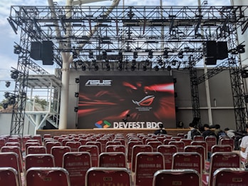 A stage setup with a large screen displaying logos for ASUS and Republic of Gamers. Rows of red chairs are arranged in front of the stage. The event is titled 'DEVFEST BDC', suggesting a tech or gaming conference. The venue appears to be indoors with a visible ceiling truss structure.