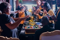 A group of people gathered around a table with a large, decorated cake and colorful spheres. Their hands are outstretched, suggesting a gesture of blessing or celebration. The setting is indoors, with festive decorations like balloons visible in the background.