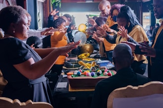 A group of people gathered around a table with a large, decorated cake and colorful spheres. Their hands are outstretched, suggesting a gesture of blessing or celebration. The setting is indoors, with festive decorations like balloons visible in the background.