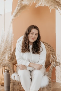 A person with long curly hair sits comfortably on a woven chair. They wear a white hoodie and pants, surrounded by pampas grass and a warm, earthy-toned backdrop. The setting suggests a cozy and natural aesthetic.