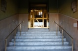 A set of marble stairs with golden handrails leading up to a doorway that appears to be made of glass and metal. The walls on either side of the stairs are painted in a muted color, and there are glass light fixtures mounted on each wall. The doorway opens into a well-lit interior space, possibly indicating a public or commercial building.