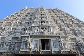 An intricately carved temple facade stretches upwards towards a clear blue sky. The architectural structure is adorned with numerous statues and reliefs of deities, showcasing detailed craftsmanship and elaborate design. Each level of the structure appears to have its own narrative, captured in stone figures that resemble traditional Indian iconography.
