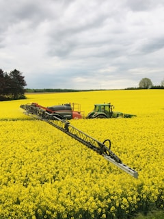 A green tractor is working in a vast field of bright yellow flowers, likely canola or rapeseed. The tractor is towing a large sprayer, applying substances over the crop. In the background, a line of trees separates the field from a cloudy sky.