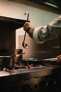 A person is cooking on a stove in a kitchen, with several pots and pans on the burners. One hand holds a ladle, while the other is handling a pan on an active flame. The setting appears to be a professional kitchen environment with stainless steel appliances.