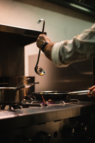 A person is cooking on a stove in a kitchen, with several pots and pans on the burners. One hand holds a ladle, while the other is handling a pan on an active flame. The setting appears to be a professional kitchen environment with stainless steel appliances.