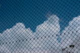 A chain-link fence with a backdrop of blue sky and fluffy white clouds, creating a contrast between the rigidity of the fence and the softness of the clouds.
