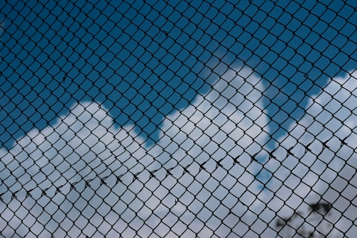 A chain-link fence with a backdrop of blue sky and fluffy white clouds, creating a contrast between the rigidity of the fence and the softness of the clouds.