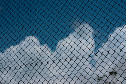 A chain-link fence with a backdrop of blue sky and fluffy white clouds, creating a contrast between the rigidity of the fence and the softness of the clouds.