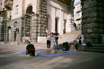 A group of young people are gathered outside a historic building with stone walls. One person in front is captured mid-movement, possibly dancing, on a wooden platform placed over a blue mat. Some are sitting nearby, and there is a large speaker next to the group, indicating an outdoor performance or practice.