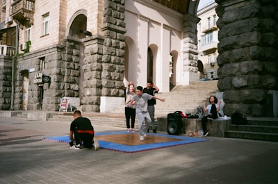 A group of young people are gathered outside a historic building with stone walls. One person in front is captured mid-movement, possibly dancing, on a wooden platform placed over a blue mat. Some are sitting nearby, and there is a large speaker next to the group, indicating an outdoor performance or practice.