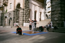 A group of young people are gathered outside a historic building with stone walls. One person in front is captured mid-movement, possibly dancing, on a wooden platform placed over a blue mat. Some are sitting nearby, and there is a large speaker next to the group, indicating an outdoor performance or practice.