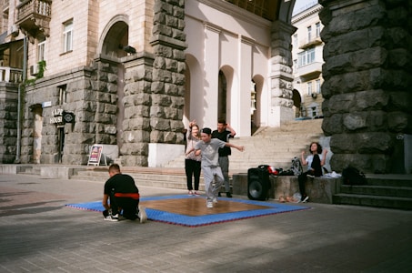 A group of young people are gathered outside a historic building with stone walls. One person in front is captured mid-movement, possibly dancing, on a wooden platform placed over a blue mat. Some are sitting nearby, and there is a large speaker next to the group, indicating an outdoor performance or practice.