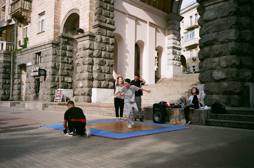 A group of young people are gathered outside a historic building with stone walls. One person in front is captured mid-movement, possibly dancing, on a wooden platform placed over a blue mat. Some are sitting nearby, and there is a large speaker next to the group, indicating an outdoor performance or practice.
