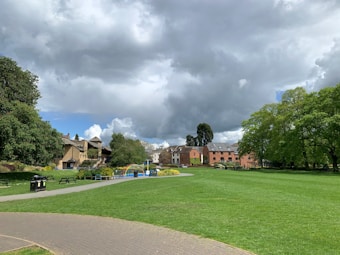 A lush green park under a cloudy sky, featuring manicured lawns, mature trees, and a few residential buildings in the background. A paved walkway winds through the grassy area, and there are benches and trash bins positioned along it. The buildings have a classic architectural design and are surrounded by landscaping with shrubs and flowers.