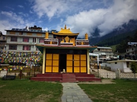 A small, colorful temple with a distinct yellow and red gabled roof sits prominently in the foreground. The temple is surrounded by a garden with grass and pathways leading up to it. Behind the temple, multicolored prayer flags are strung along a fence, adding vibrancy to the scene. Residential buildings with a more modern architectural style appear in the background, situated against a backdrop of misty mountains and pine trees under a partly cloudy sky.