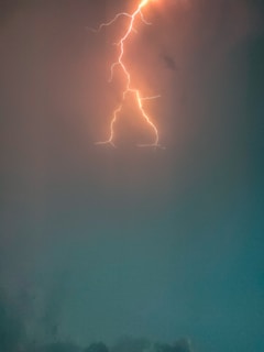 A vibrant lightning bolt streaks across a darkened sky, illuminating the clouds with orange and yellow hues. The surrounding atmosphere has a mix of darker and lighter shades of blue and gray.