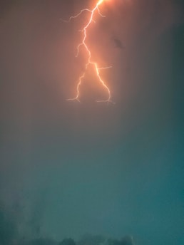 A vibrant lightning bolt streaks across a darkened sky, illuminating the clouds with orange and yellow hues. The surrounding atmosphere has a mix of darker and lighter shades of blue and gray.