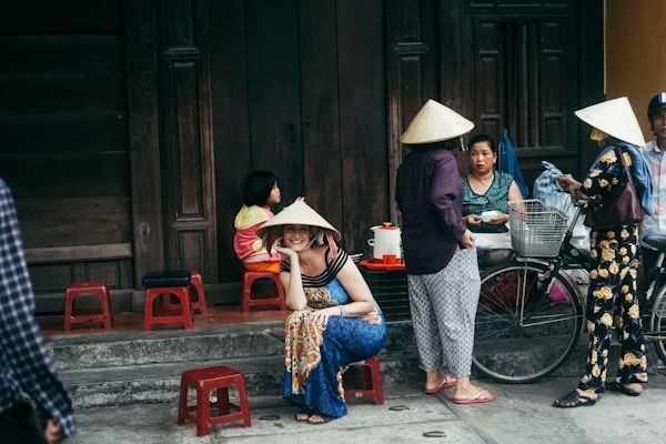 A group of people wearing traditional conical hats are gathered in a street setting. One person is sitting on a small stool, smiling, while others stand around engaged in conversation. A child is sitting nearby with bright clothing. The scene includes a wooden door in the background and a bicycle with bags attached.