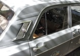 A classic car with a sleek, vintage design, featuring a distinctive side emblem and chrome detailing. The vehicle's body is a muted green color with reflective metal elements. The interior is visible through the windows, showing brown leather seats and a steering wheel. Sunlight creates dappled shadows across the car's surface.