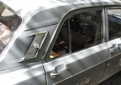 A classic car with a sleek, vintage design, featuring a distinctive side emblem and chrome detailing. The vehicle's body is a muted green color with reflective metal elements. The interior is visible through the windows, showing brown leather seats and a steering wheel. Sunlight creates dappled shadows across the car's surface.