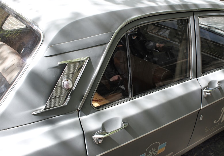 A classic car with a sleek, vintage design, featuring a distinctive side emblem and chrome detailing. The vehicle's body is a muted green color with reflective metal elements. The interior is visible through the windows, showing brown leather seats and a steering wheel. Sunlight creates dappled shadows across the car's surface.