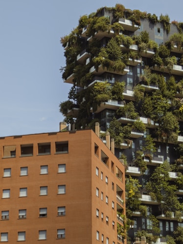 A modern building with an innovative design incorporating greenery and trees on its balconies is juxtaposed with a traditional brick apartment building. The vertical garden creates a striking contrast with the red-bricked structure.