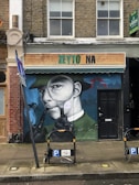 A mural on a brick building features a person wearing a suit and hat, holding a pipe in their mouth and looking through a magnifying glass. The shop sign above reads 'ZEYTO NA' and an adjacent 'To Let' sign is visible. Two bicycle parking stands are in front of the mural.