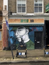 A mural on a brick building features a person wearing a suit and hat, holding a pipe in their mouth and looking through a magnifying glass. The shop sign above reads 'ZEYTO NA' and an adjacent 'To Let' sign is visible. Two bicycle parking stands are in front of the mural.