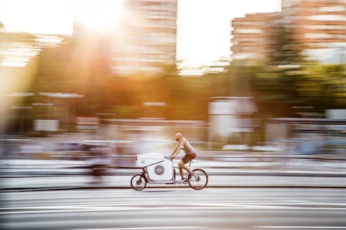 A cyclist rides a cargo bike swiftly along a city street with buildings and trees blurred in the background, suggesting movement and speed. Sunlight streams through, adding a warm glow to the scene.