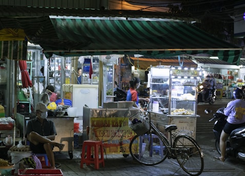 A bustling street food market scene at night, featuring several stalls with vendors and customers. One vendor is preparing food behind a brightly lit display, while a person sits on a red plastic stool nearby. A bicycle is parked in front, and the awnings of the stalls display striped patterns. The atmosphere is lively with various lights, people, and activities.