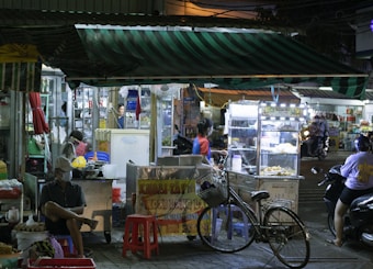 A bustling street food market scene at night, featuring several stalls with vendors and customers. One vendor is preparing food behind a brightly lit display, while a person sits on a red plastic stool nearby. A bicycle is parked in front, and the awnings of the stalls display striped patterns. The atmosphere is lively with various lights, people, and activities.