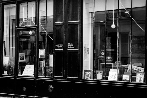 A vintage storefront with large glass windows displaying various books and posters. The signage on the door includes phrases in French, suggesting a bookshop or literary establishment. The overall appearance is classic and timeless.