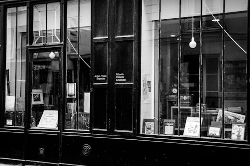 A vintage storefront with large glass windows displaying various books and posters. The signage on the door includes phrases in French, suggesting a bookshop or literary establishment. The overall appearance is classic and timeless.