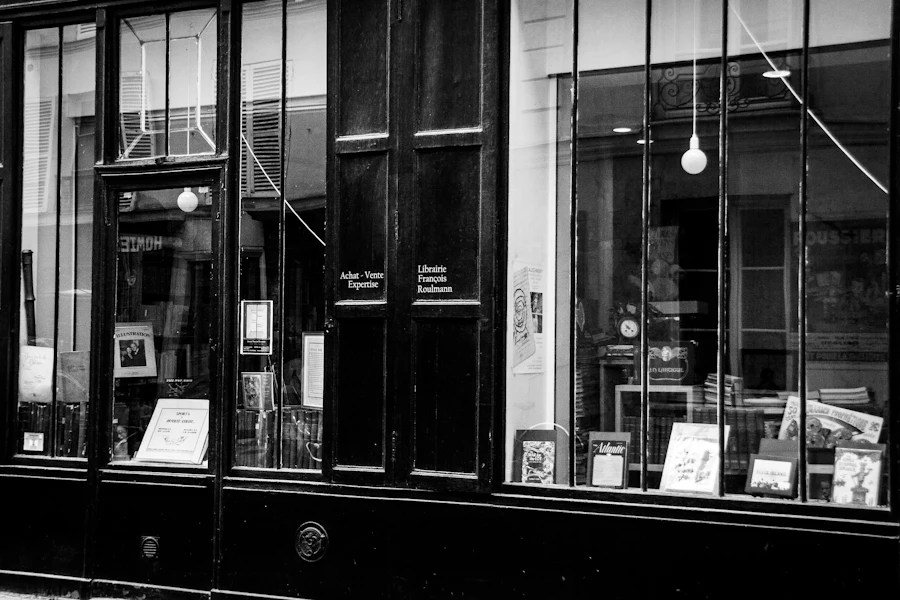 Black and white photograph of a dark timber shopfront, its tall windows stacked with framed prints and books under two bare hanging bulbs.