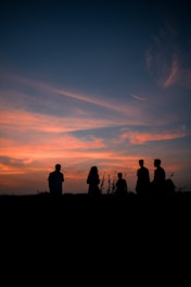 Silhouettes of five people stand against a vibrant sunset sky with orange and blue hues, creating a serene and peaceful scene.