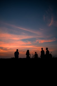 Silhouettes of five people stand against a vibrant sunset sky with orange and blue hues, creating a serene and peaceful scene.