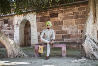 A person wearing a light-colored kurta and a green turban is sitting on a wooden bench. The setting is outdoors by an old stone building with archways and a tree with exposed roots.