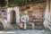 A person wearing a light-colored kurta and a green turban is sitting on a wooden bench. The setting is outdoors by an old stone building with archways and a tree with exposed roots.