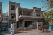 A two-story building is under construction, featuring a concrete structure with unfinished walls and open windows. Construction materials such as sand, bricks, and a stack of bags are visible in the front yard, along with some makeshift scaffolding. There is a blue barrel on the ground and some greenery on the right side of the image.